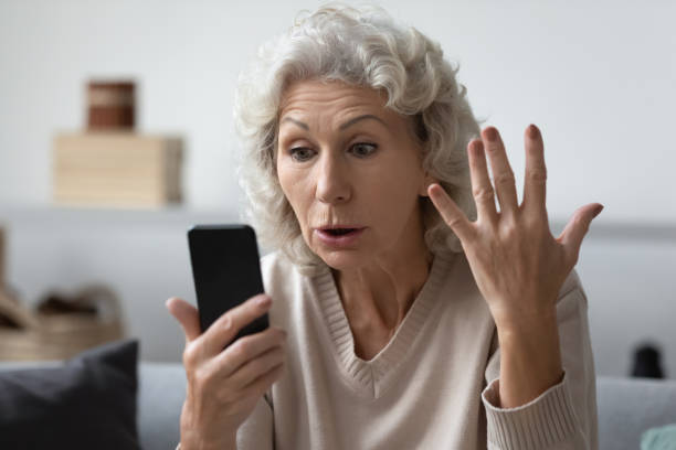 Angry shocked mature phone user staring at screen in surprise Angry shocked mature phone user staring at screen in surprise. Elderly lady worried about app error, data stealing, scam, bad news, problems with smartphone. Annoyed woman using cell for video call scammer stock pictures, royalty-free photos & images