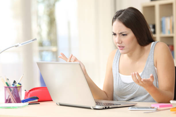 Angry student looking at laptop on a desk at home Angry student looking at laptop on a desk at home college scams online stock pictures, royalty-free photos & images