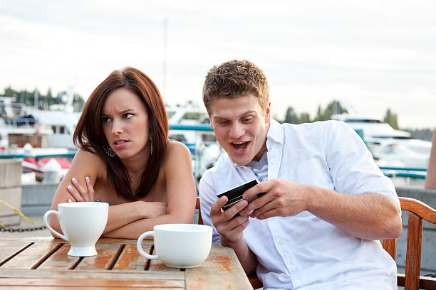 Annoying Boyfriend with Smart Phone Photo a young couple having a disasterous first date.  Young woman looks annoyed as her date seems more excited about his phone than he is about being with her. bad date stock pictures, royalty-free photos & images