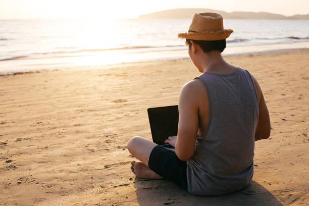 Anonymous freelancer using laptop near sea Back view of barefoot man in hat sitting on sandy beach near waving sea and browsing modern laptop nomadic people stock pictures, royalty-free photos & images