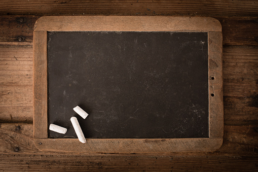 Color stock photo of an old, well-used school slate chalkboard with wood frame and chalk sitting on an old wooden trunk.