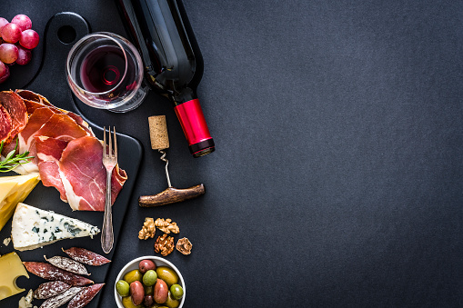 Appetizer: top view of a black background with a composition of red wine bottle, wineglass, a cutting board with various cheeses and Iberico ham arranged at the left border leaving useful copy space for text and/or logo at the right. Some grapes, olives, nuts and a corkscrew complete the composition. Predominant colors are red and black. XXXL 42Mp studio photo taken with Sony A7rii and Sony FE 90mm f2.8 macro G OSS lens