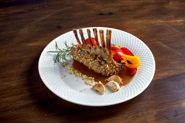 Appetizing roasted rack of lamb in nuts, with sauce and grilled vegetables, served in a white plate on a wooden background. stock photo