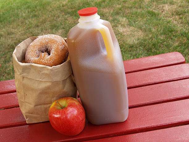 apple cider and donuts apple cider, an apple and a bag of cinnamon-sugared donuts on a red picnic table cider stock pictures, royalty-free photos & images