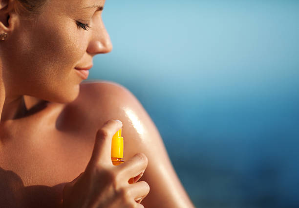 Applying suntan lotion. Closeup of adult caucasian woman spraying suntan lotion onto her shoulder. It's sunny summer day and she's sunbathing with a smirk on her face. Blurry sea in background. sunscreen stock pictures, royalty-free photos & images