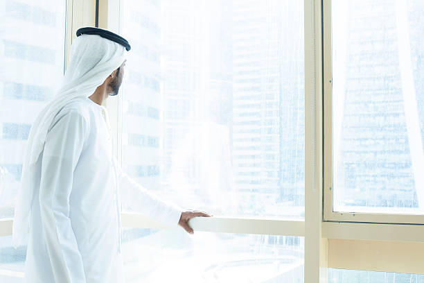 Arab businessman looking out of a business building window Arab businessman looking through business building window. Businessman is  worn in traditional male dress  kandura (dishdasha) with  kaffiyeh (white headdress of cotton fabric) held in place by a rope (agal). Concept for modern business in Middle East.  Image is taken during Dubai Istockalypse in United Arab Emirates. agal stock pictures, royalty-free photos & images