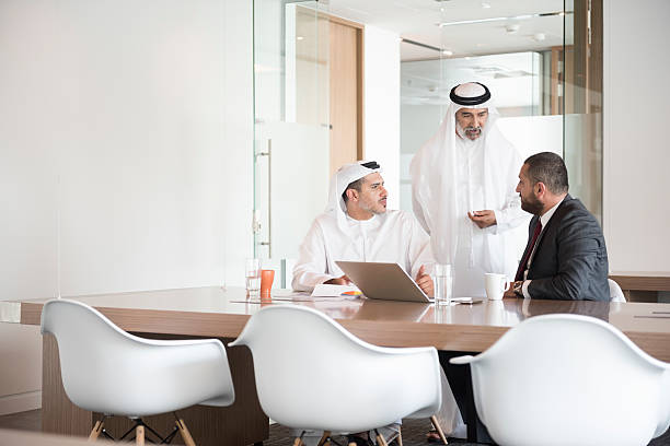 Arab businessmen discussing at conference table A photo of serious Middle Eastern businessmen discussing. Traditional Arab Emirati men in thobe talk to man in western suit. Middle East professional business people are in a meeting with laptop at conference table, Dubai, United Arab Emirates. agal stock pictures, royalty-free photos & images