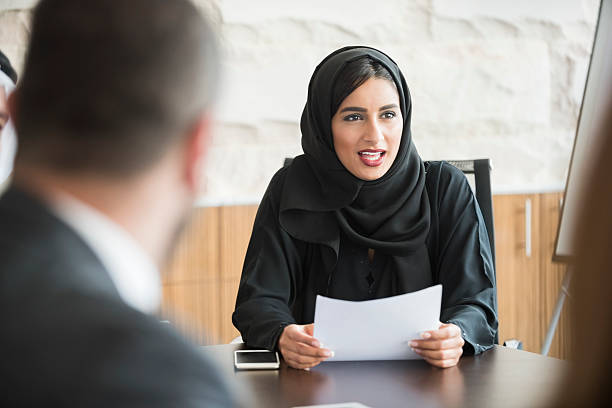 Arab businesswoman wearing traditional abaya in business meeting A photo of mature businessman looking at colleague. He is wearing traditional clothes. Professional is sitting and smiling at coworkers. Office workers are in a meeting, at brightly lit workplace. middle eastern culture stock pictures, royalty-free photos & images