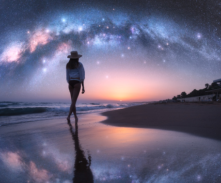 Arched Milky Way and young woman in hat on sandy beach against starry sky reflected in water at night. Landscape. Summer travel. Girl on the tropical sea coast and bright Milky Way arch, stars. Space
