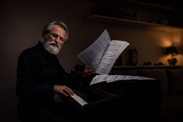 Arranged gray-haired, bearded, charming composer, pianist, middle-aged artist in a black shirt at home in a darkened room with ambient lamp and light, concentrated, stacking and playing the electric piano in between, drinking a cup of tea or coffee. stock photo