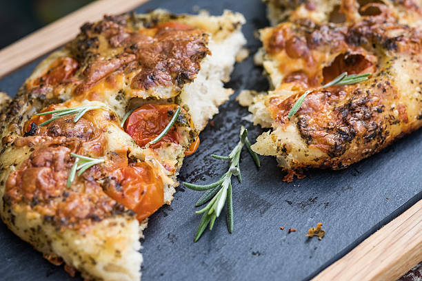 Artisan Focaccia Bread Pizza with Cherry Tomatoes, Pesto, Rosemary Vegetarian pizza.  Close up of a hand made focaccia bread topped with cherry tomatoes, pesto, cheese and herbs served on a slate serving platter.  Torn in half and garnished with fresh rosemary. artisanal food and drink stock pictures, royalty-free photos & images