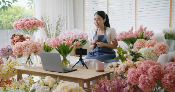Asia girl vlogger influencer or SME owner people smile work on home video camera selfie shoot filming for live sell show happy talk on mobile VoIP app. Remote sale product at modern florist gift shop. stock photo
