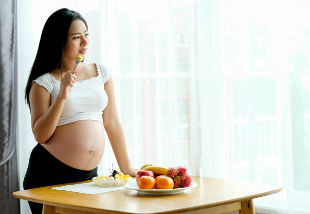 người phụ nữ mang thai xinh đẹp châu á đứng và ăn một số loại trái cây trước cửa sổ kính với rèm trắng - mang thai hình ảnh sẵn có, bức ảnh & hình ảnh trả phí bản quyền một lần