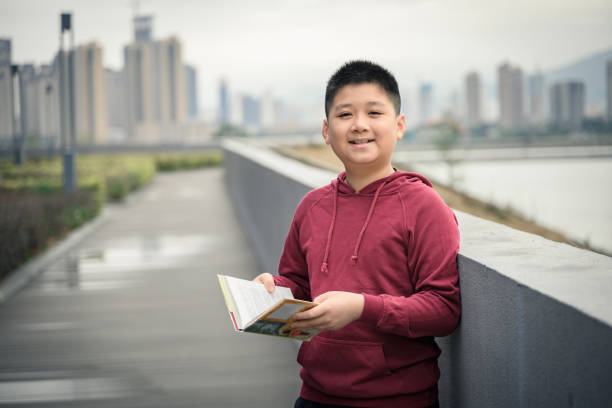 Asian boy reads a book against the fence in the park. stock photo