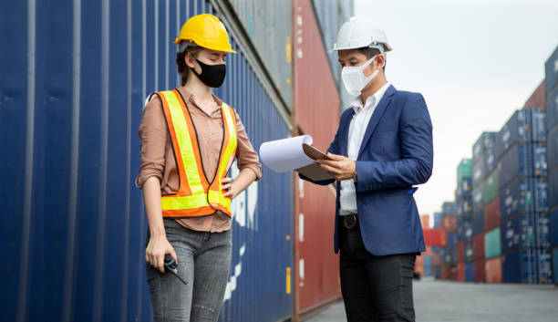 asian businessman using clipboard and harbor worker woman  wareing face mask checking list inventory loading Containers box at warehouse logistic in Cargo during coronavirus, covid 19 pandemic stock photo