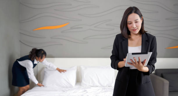 Asian businesswoman in black suit standing with tablet computer in her hand. The hotel manager verify the tidiness of the room for hotel guests while the maid making the bed in the background. stock photo