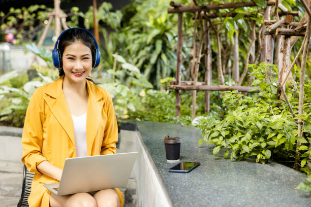 Asian businesswoman sitting in Bluetooth headphones working on laptop outdoors happy smile. lady using tablet happily public park. use of modern internet communication technology. white collar worker stock photo