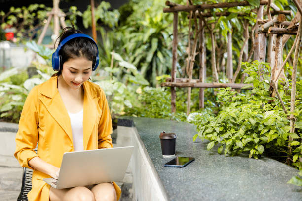 Asian businesswoman sitting in Bluetooth headphones working on laptop outdoors happy smile. lady using tablet happily public park. use of modern internet communication technology. white collar worker stock photo