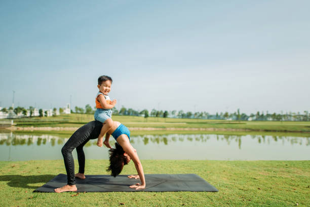 Asian chinese beautiful women practicing yoga with her son at public lake park wide shot of child looking at camera with smiling face and sitting on mother tummy while mother do stretching exercise yoga pose on yoga mat outdoor asian yoga pants stock pictures, royalty-free photos & images