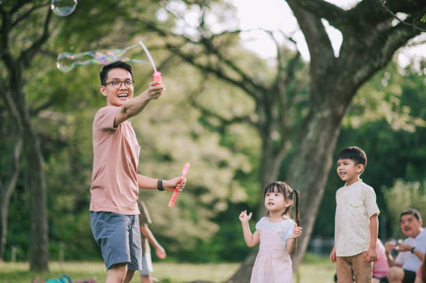 asian chinese father playing with his children with bubble wand at public park in the morning backlit sunlight asian chinese father playing with his children with bubble wand at public park in the morning backlit sunlight fathers day stock pictures, royalty-free photos & images
