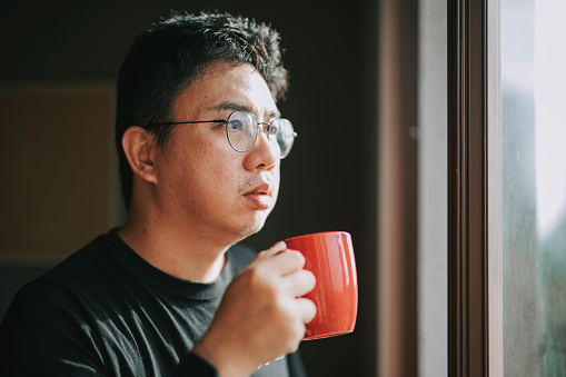Asian chinese mid adult man  looking outside of window from his home during sunset with serious facial expression holding a hot drink mug