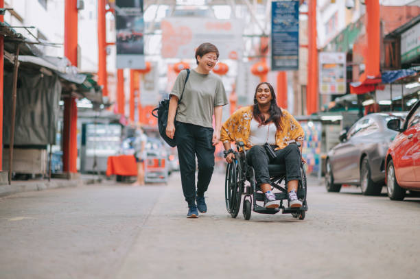 asian chinese mid adult woman walking side by side with her indian female friend with disability on wheelchair at city street - wheelchair street happy imagens e fotografias de stock
