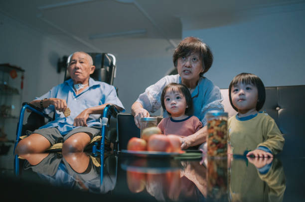 asian chinese senior couple with wheelchair and twin grandson enjoying movie  night at home watching tv in living room sitting on sofa asian chinese senior couple with wheelchair and twin grandson enjoying movie  night at home watching tv in living room sitting on sofa asian kids watching tv stock pictures, royalty-free photos & images