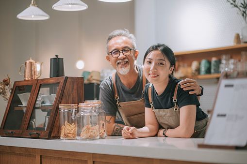 asian chinese senior male cafe owner and her daughters looking at camera smiling at coffee shop counter