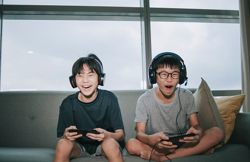 2 asian chinese sibling brother sitting on sofa playing online video games with headset in living room during weekend