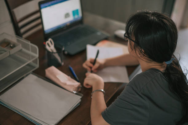 asian chinese teenage girl doing homework studying at home in front of laptop in kitchen asian chinese teenage girl doing homework studying at home in front of laptop in kitchen distance learning stock pictures, royalty-free photos & images