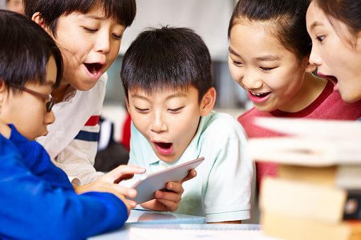group of asian elementary school children gathering around playing game together using tablet during break.