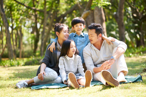 asian family sitting on grass talking chatting asian family with two children having fun sitting on grass talking chatting outdoors in park asian mother talking with daughter stock pictures, royalty-free photos & images