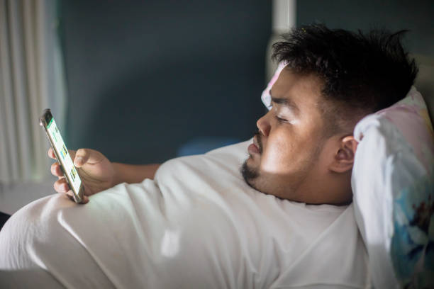 Asian fat man using a phone before sleep Close up of an Asian fat man lying on the bed while using a mobile phone before sleep at night time fat man looks at the phone stock pictures, royalty-free photos & images