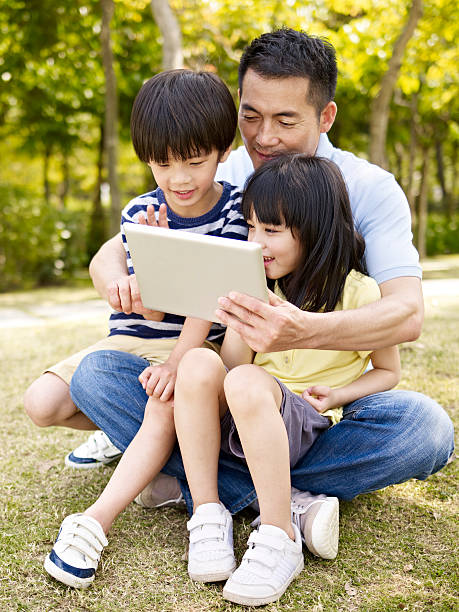 asian father and children using tablet outdoors asian father and two children sitting on grass looking at tablet computer, outdoor in a park. child korea little girls korean ethnicity stock pictures, royalty-free photos & images