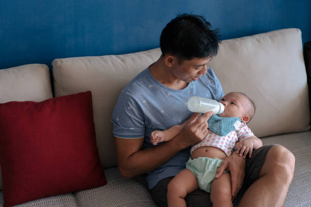 Asian father bottle feeding his toddler daughter on a couch stock photo