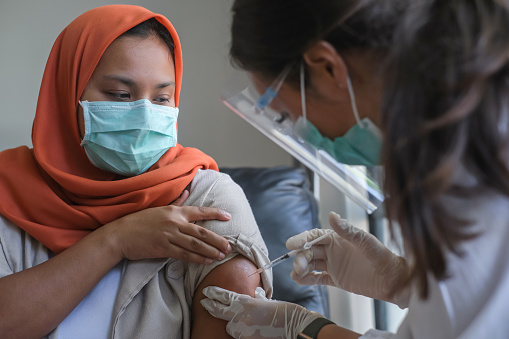 Close up shot of Asian female doctor giving a muslim patient the shot of vaccine. The doctor wearing a face shield, surgical mask and surgical gloves for safety protection.