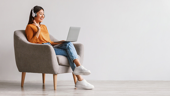 Full length of young Asian female in headphones using laptop for online communication, video chatting, waving at webcam, sitting in armchair against white studio wall, banner design with copy space