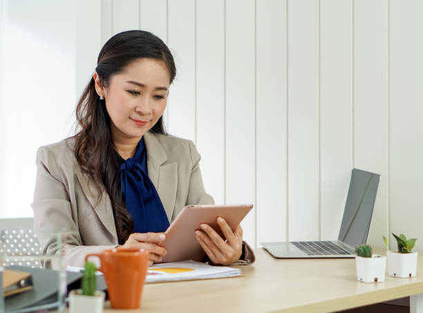 Asian female office manager with brown suit holding tablet computer in her hand. Morning work atmosphere in a modern office. stock photo
