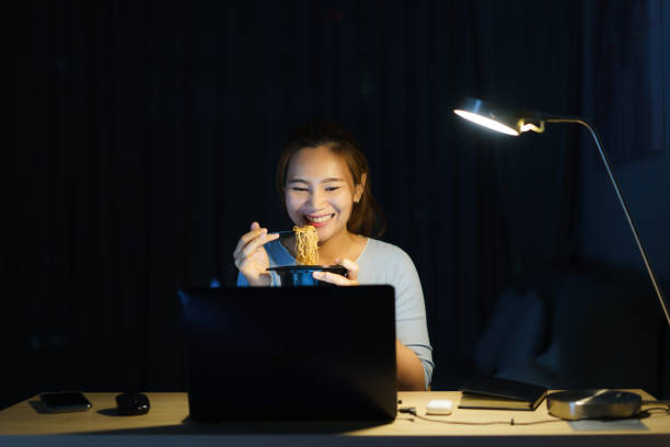 Asian freelancer smart business woman eating instant noodles while working on a laptop in the living room at home at night happy Asian girl sitting on a desk overtime enjoying relaxing time stock photo