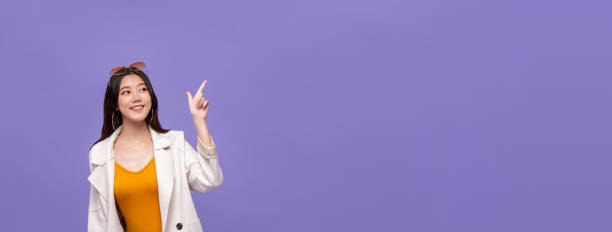 Asian girl pointing hand to empty space on purple banner background Young pretty smiling Asian woman in smart casual clothes pointing hand to empty space aside on purple banner background cute thai girl stock pictures, royalty-free photos & images