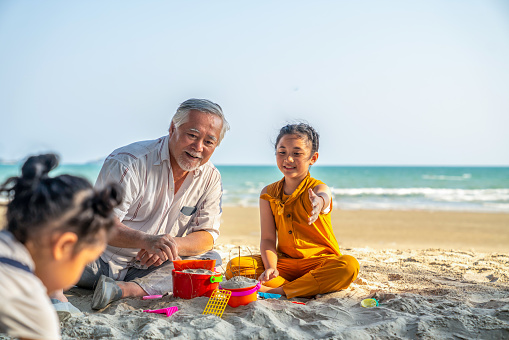 Happy multi-generation Asian family on summer vacation. Grandfather play beach toys with two little grandchild girl on the beach in sunny day. Retirement senior man with granddaughter enjoy and having fun outdoor lifestyle activity.