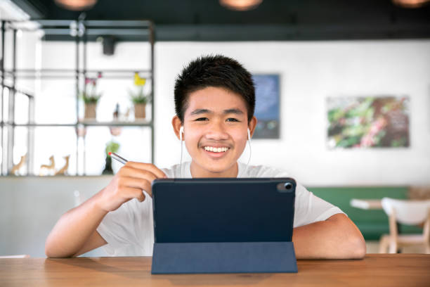 asian happy boy student wear headphone and study online with video call teached by teacher with smart tablet and laptop from home during covid-19 stock photo