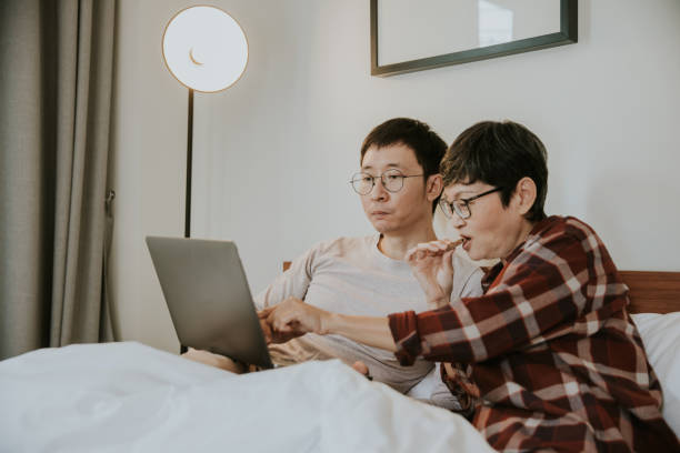Asian happy son and mother lay down in bed and wathing clips in laptop together on weekend - stock photo stock photo