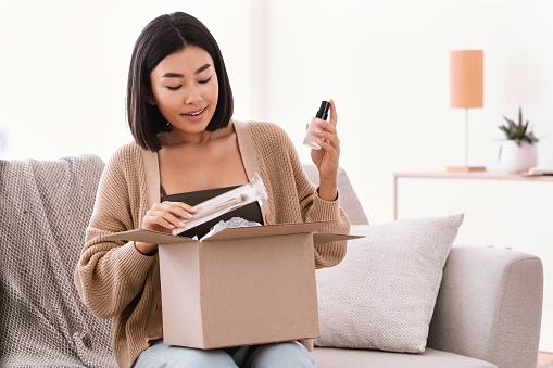 Online Shopping. Smiling asian woman opening parcel box with cosmetics at home, holding makeup products, unpacking gift sitting on the couch at home. Lady satisfied with delivery service