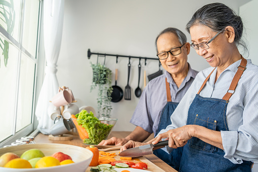 Asian loving senior elderly couple wear apron and cooking in kitchen. Attractive strong old man and woman grandparent wear eyeglass enjoy retirement life activity at home. Family relationship concept.