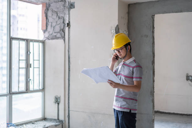 Asian male engineer using phone and working with construction drawings at renovation sites stock photo