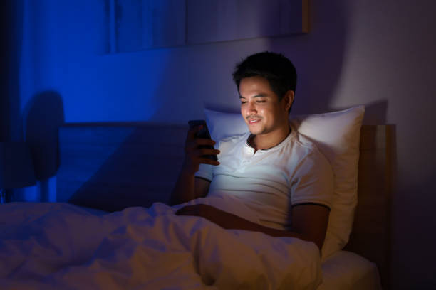 Asian man are typing online chat with a friend or girlfriend at night on a bed in a bedroom that is off the lights. stock photo