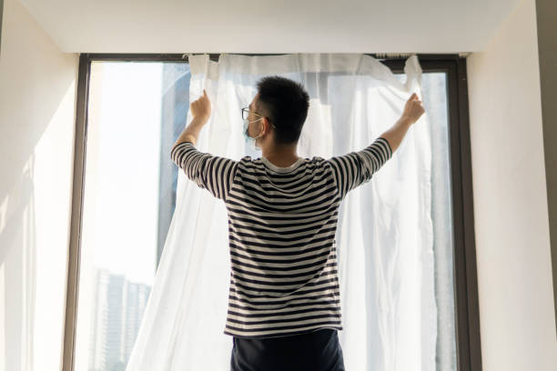 Asian man hanging curtains with protective face mask  while moving house stock photo