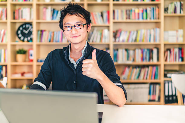 Asian man with laptop, thumbs up, at home office, library Young adult Asian man with laptop, thumbs up ok sign, home office or library scene, bookshelf with clock blur background with copy space, success or technology concept business thumbs up stock pictures, royalty-free photos & images