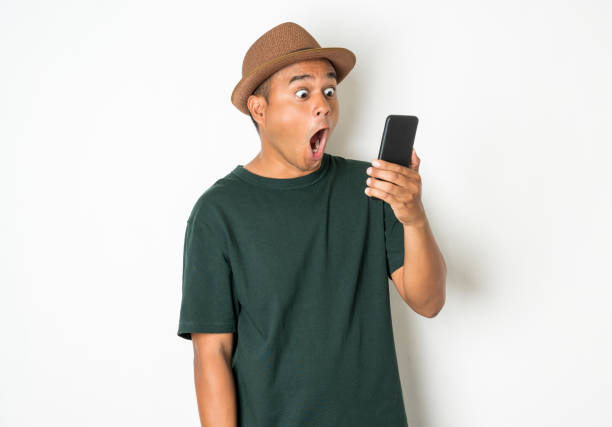 Asian men are using smartphones. He looks at the phone Then made a very surprised face He wears an old green shirt and wears a brown hat. He is around 27-35 years old. stock photo
